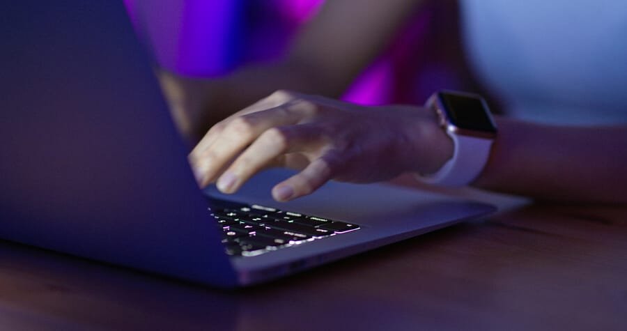 Puntuación seo de mi web beneficios de optimizar - Manos tecleando en un portátil iluminado en un ambiente con luces púrpuras y azules, con un reloj inteligente en la muñeca