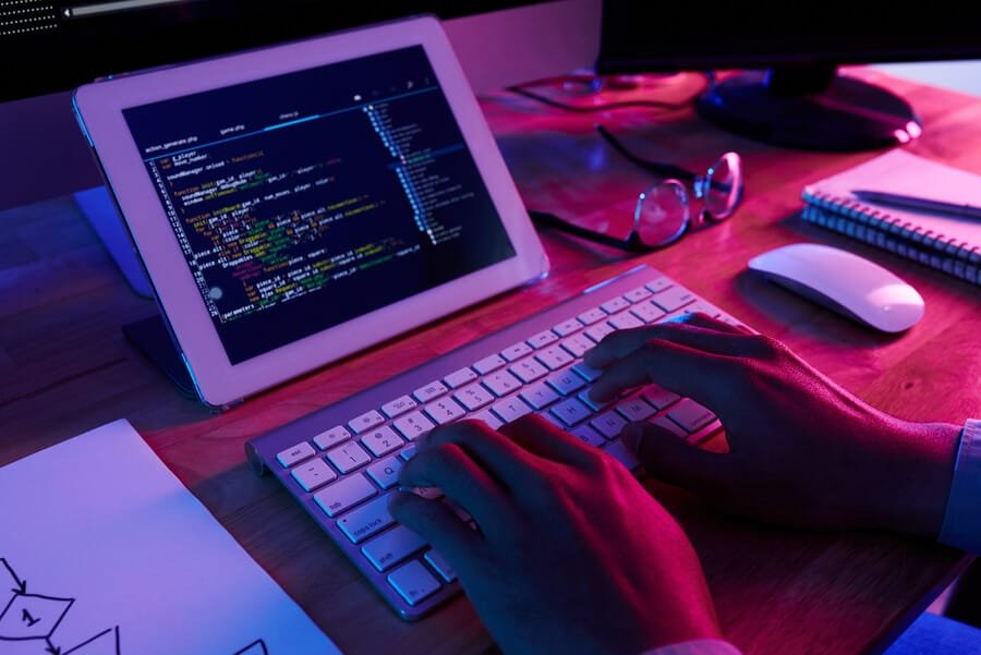 Puntuación seo de mi web estrategias para mejorar - Manos escribiendo en un teclado frente a una tableta que muestra líneas de código, en un ambiente de trabajo con iluminación tenue en tonos púrpura y azu